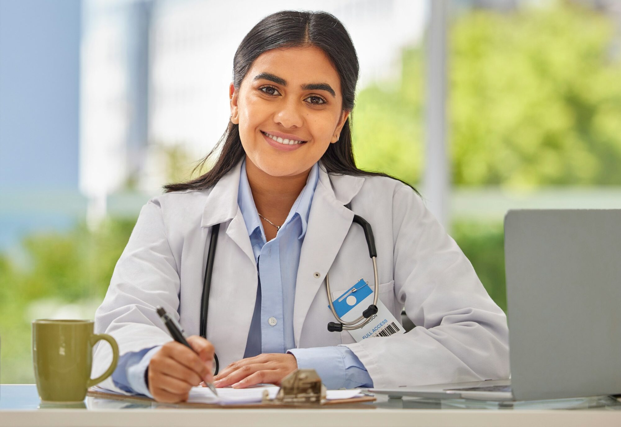Portrait of a young indian doctor wearing a stethoscope sitting in a office writing a prescription